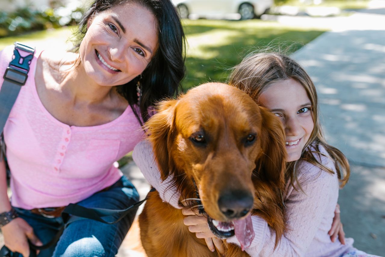 A smiling mother and daughter hugging their golden retriever on a sunny front lawn.
